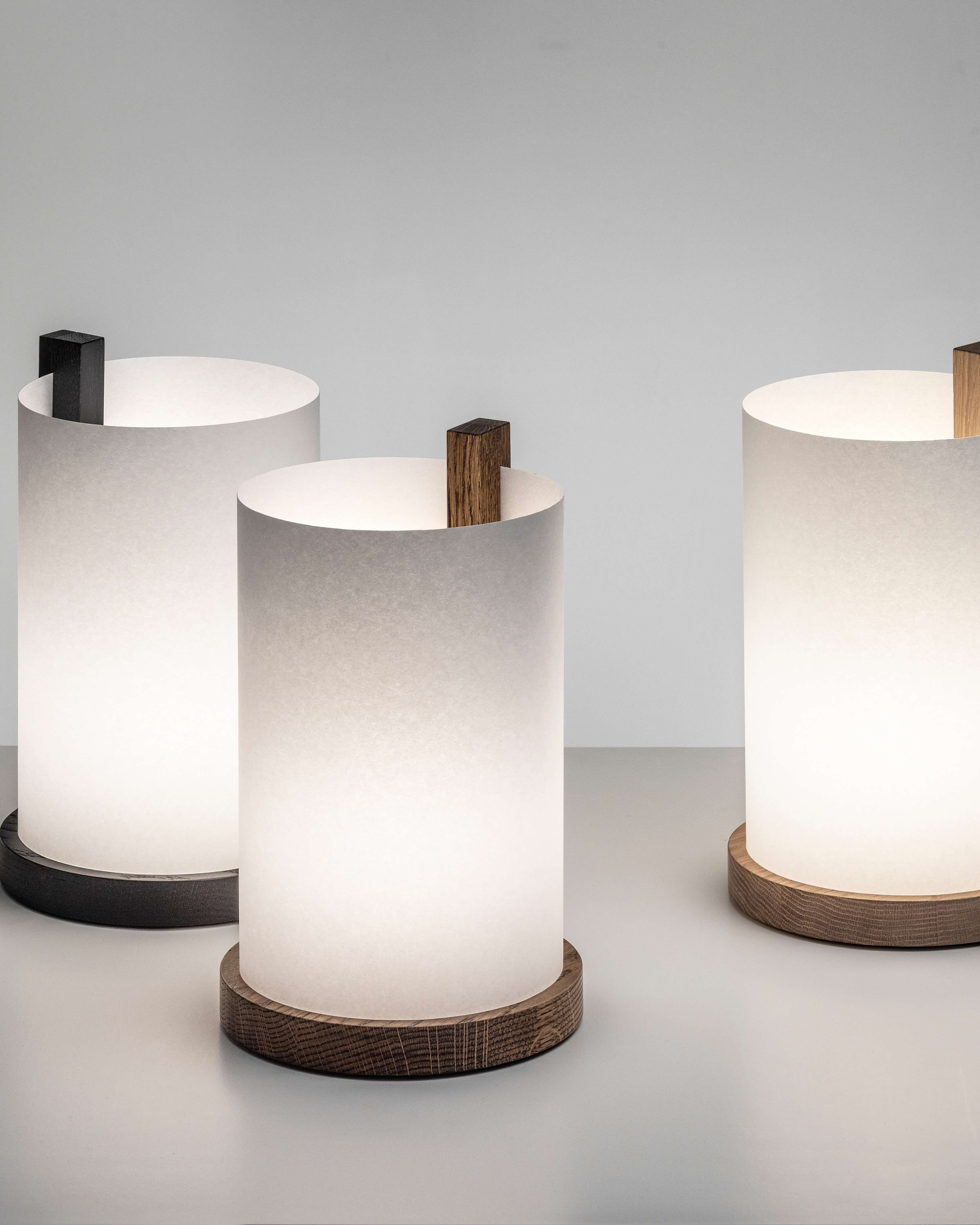 Enso table lamp in washi paper and oak with light on, three lamps with three finishes styled together on grey with background by Motarasu