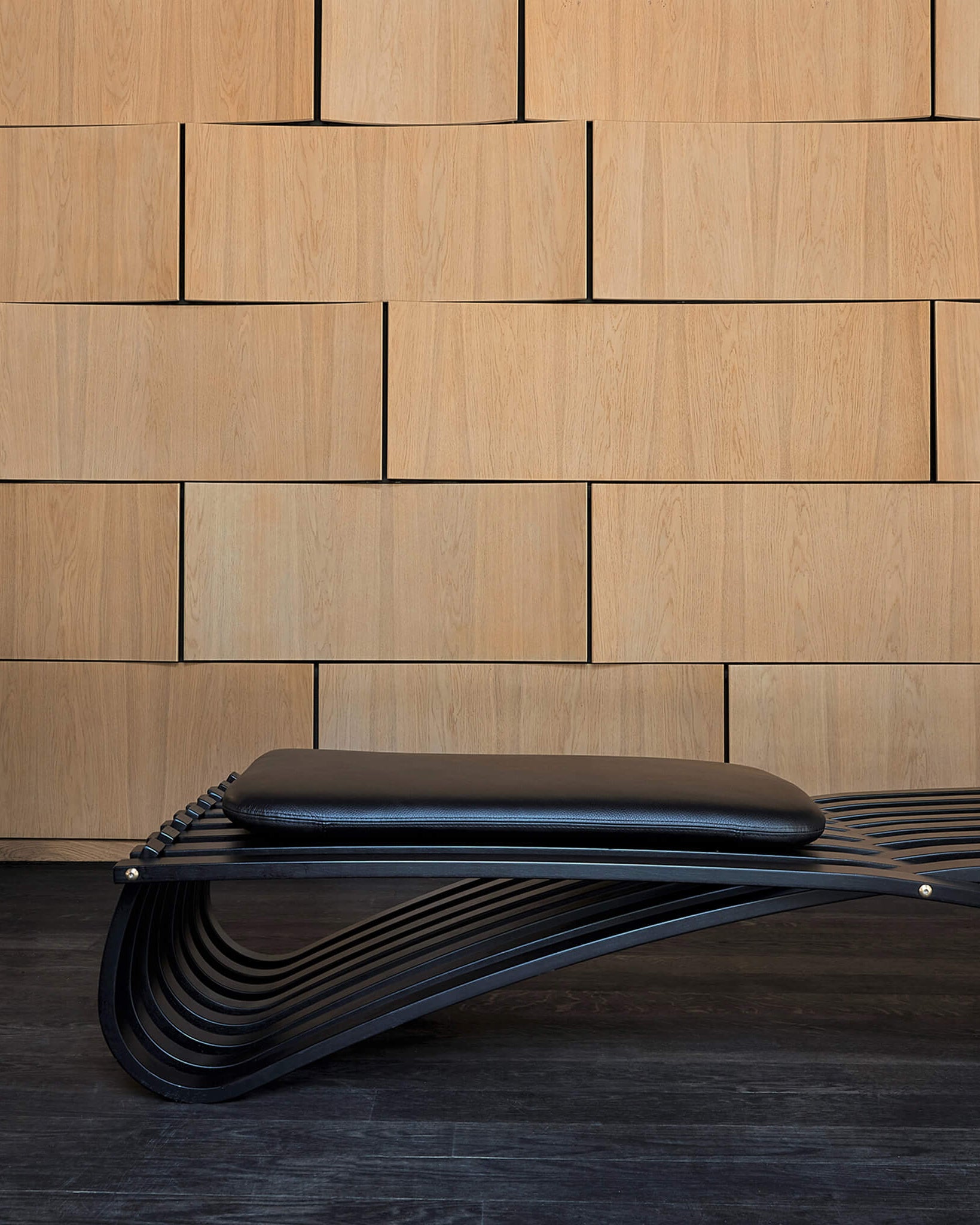 Jundo black daybed with one leather cushion, placed against a wooden wall by Motarasu