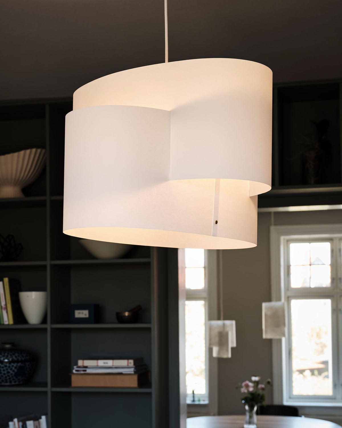 A modern cylindrical pendant light with a white washi shade, shown hanging in a dimly lit room with dark shelving and windows in the background.