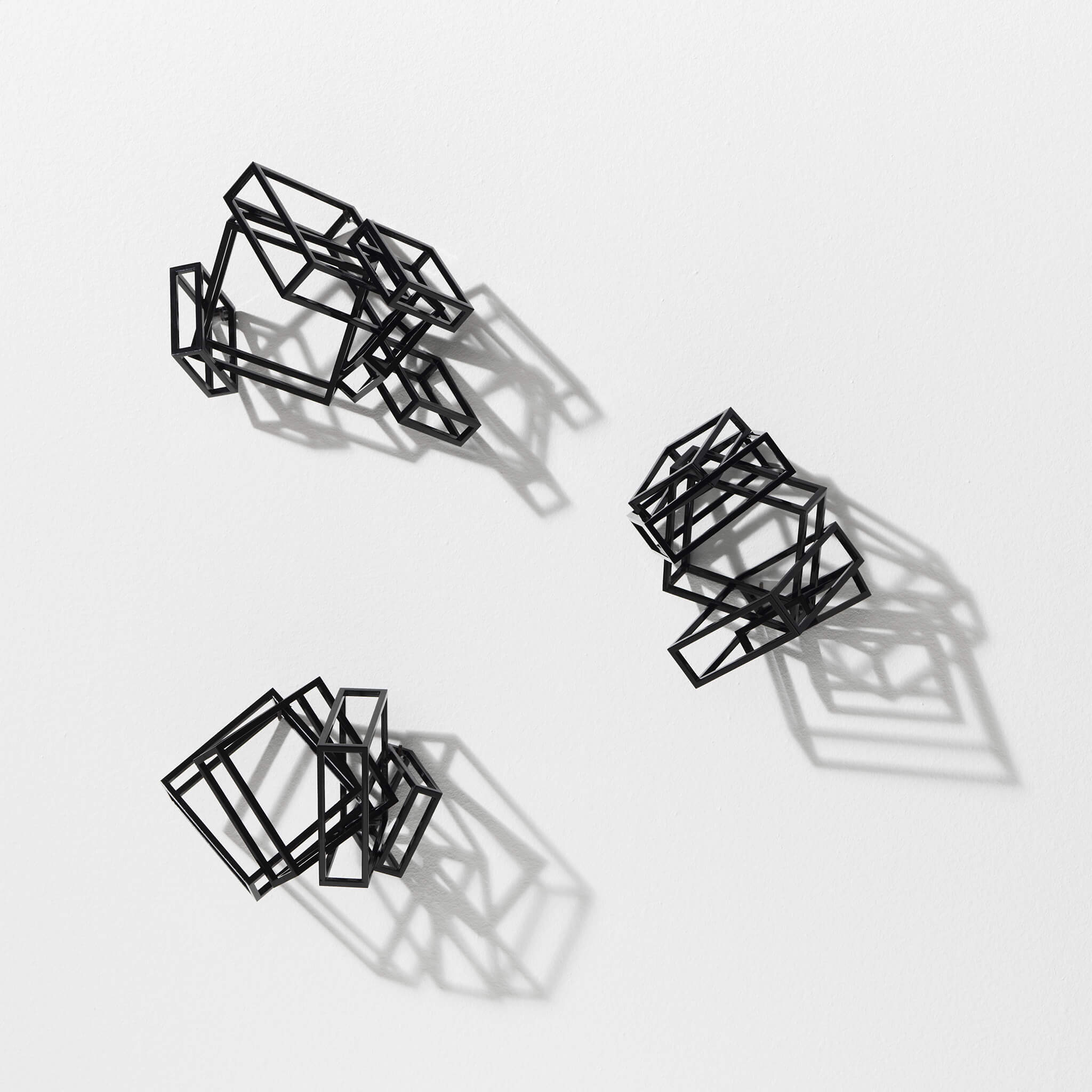 Three black coated, steel sculptures hanging on a wall and casting their shadows