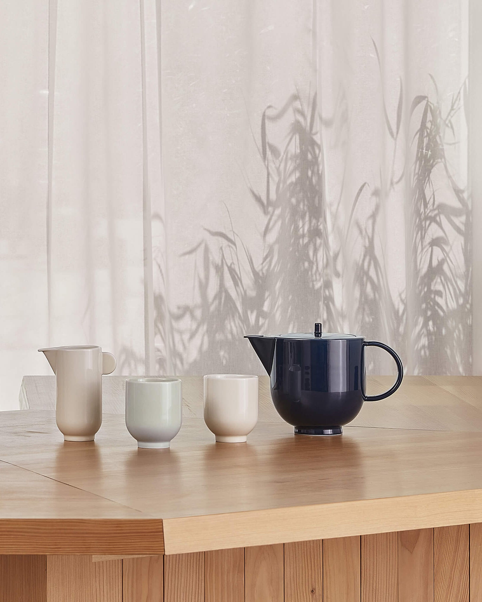 Yoko teapot in dark navy blue porcelain, styled pitcher and two mugs on wooden table side view by Motarasu