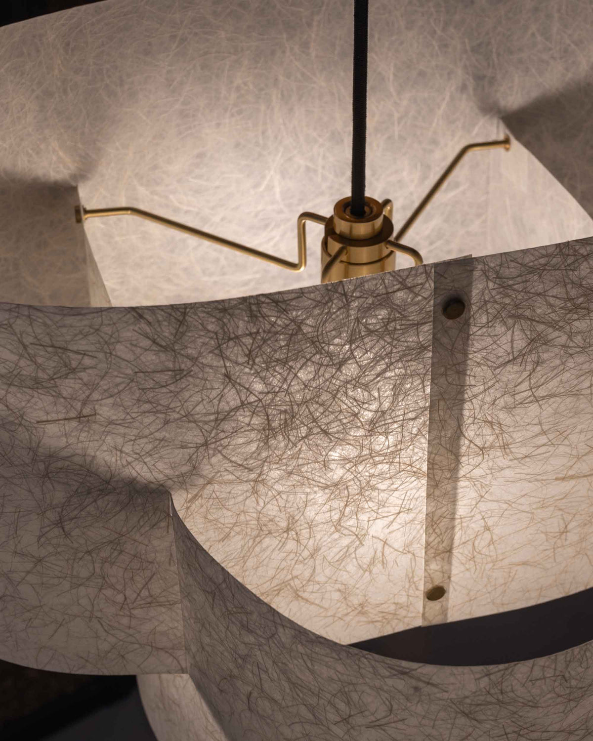 Close-up of a hanging pendant light with a textured, translucent paper shade made of fine thread-like material, illuminated from within, showing a brass fixture and black cord.