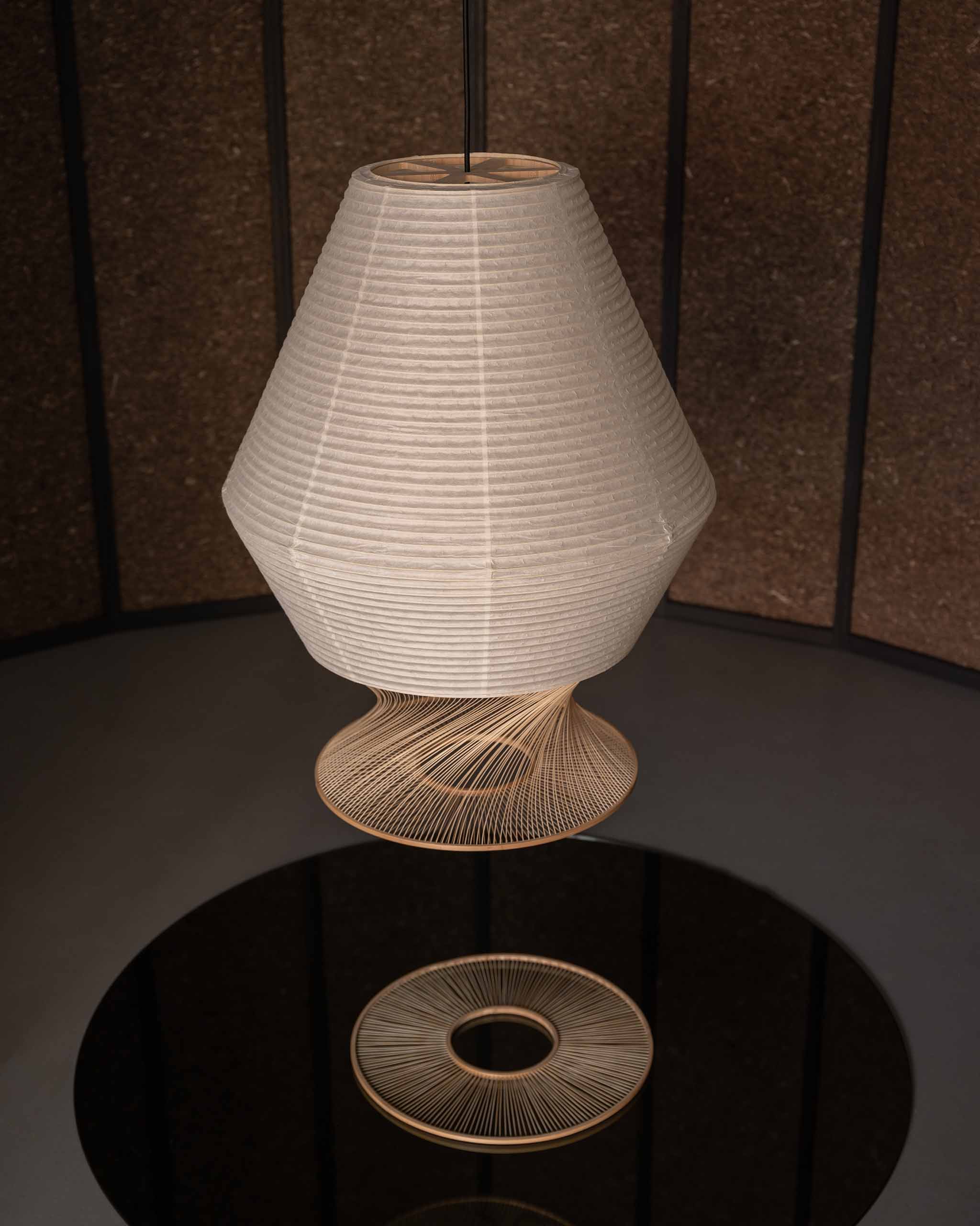 A modern pendant light with a tapered, ribbed beige fabric shade and a sculptural bamboo wire base, suspended over a reflective black table.
